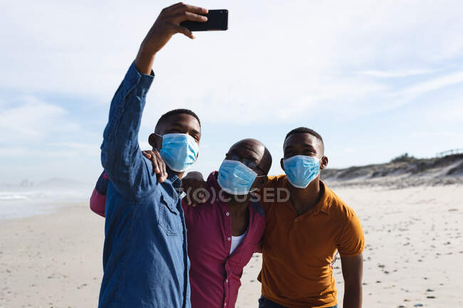 Il padre afroamericano e i suoi due figli indossano maschere facciali mentre si fanno un selfie insieme in spiaggia. regole di vacanza al mare estate durante covid-19 concetto pandemico. — Foto stock