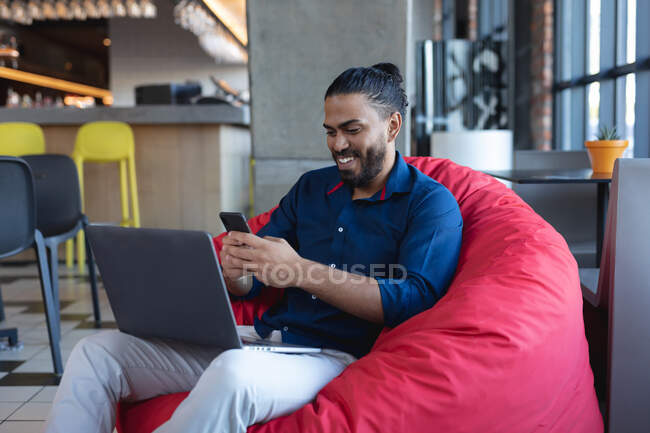 Смолящий смешанную расу мужчина сидит, используя смартфон и ноутбук в кафе. цифровые креативы на ходу. — стоковое фото