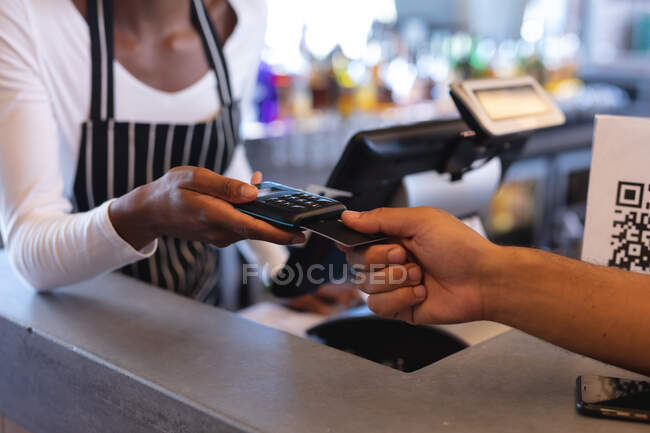 Midsection of african american waitress wearing apron, using payment terminal in cafe. independent cafe, small successful business. — Stock Photo