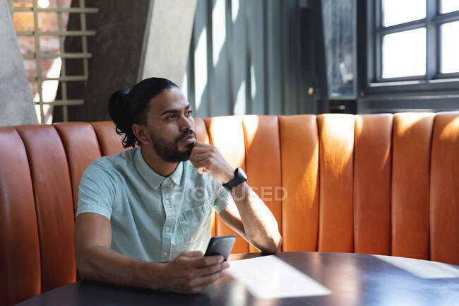 Hombre de raza mixta sentado en la cafetería, pensando y utilizando el teléfono inteligente. creativos digitales sobre la marcha. - foto de stock