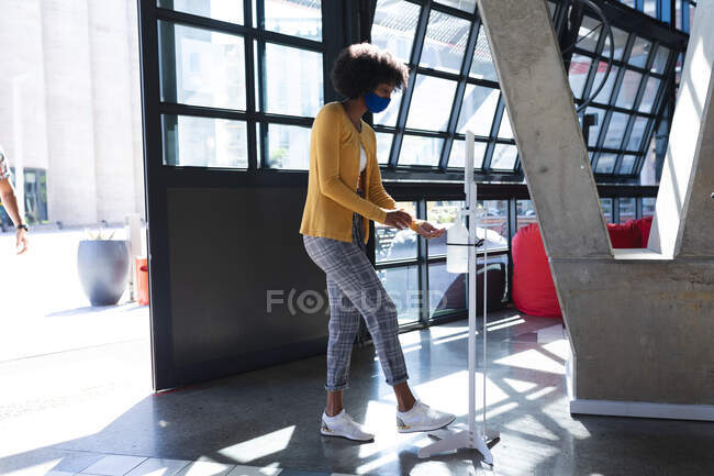 African american woman wearing face mask disinfecting hands. digital creatives on the go during coronavirus covid 19 pandemic. — Stock Photo