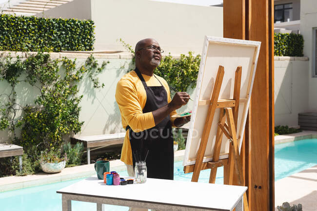 Afro-americano idoso vestindo avental pintando na tela perto da piscina. aposentadoria estilo de vida sênior conceito — Fotografia de Stock