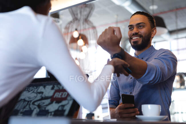 Улыбающийся клиент смешанной расы платит умными часами африканской баристе. независимое кафе, небольшой успешный бизнес. — стоковое фото
