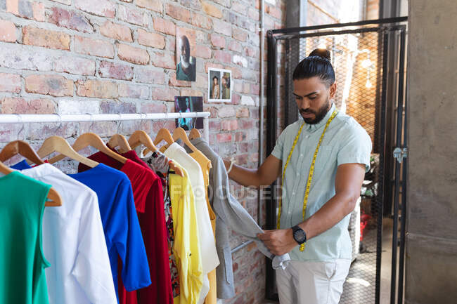 Designer masculino de raça mista usando medidor de alfaiate, empilhando roupas em cabides. negócio de design de moda criativo independente. — Fotografia de Stock