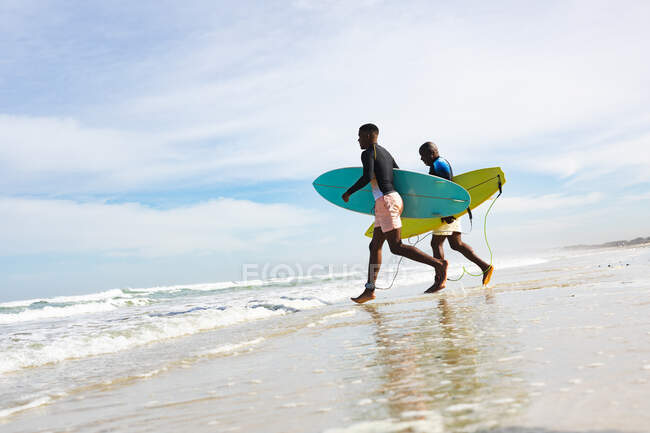 African american father and son with surfboards running towards the waves at the beach. summer beach holiday and leisure concept. — Stock Photo