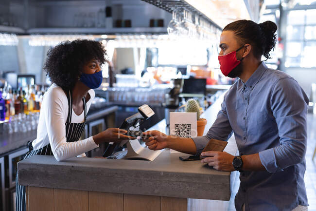 Diversos camareros masculinos y femeninos con máscaras faciales, utilizando terminal de pago en la cafetería. café independiente, negocio durante coronavirus covid 19 pandemia. - foto de stock