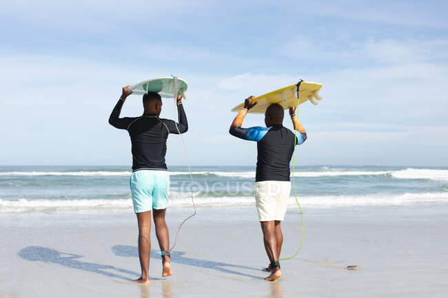 Vista trasera del padre y el hijo afroamericanos llevando tablas de surf en sus cabezas en la playa. vacaciones de playa de verano y concepto de ocio. - foto de stock
