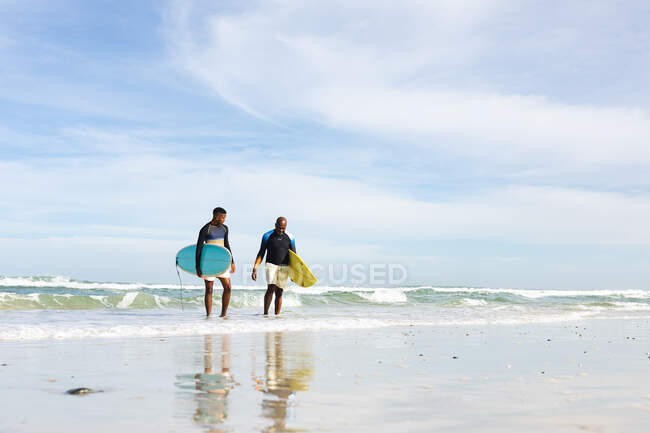 African american father and son with surfboards walking towards the beach. summer beach holiday and leisure concept. — Stock Photo