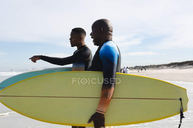 Afroamerikanischer Vater und Sohn laufen mit Surfbrettern den Wellen am Strand entgegen. Sommer-Strandurlaub und Freizeitkonzept. — Stockfoto