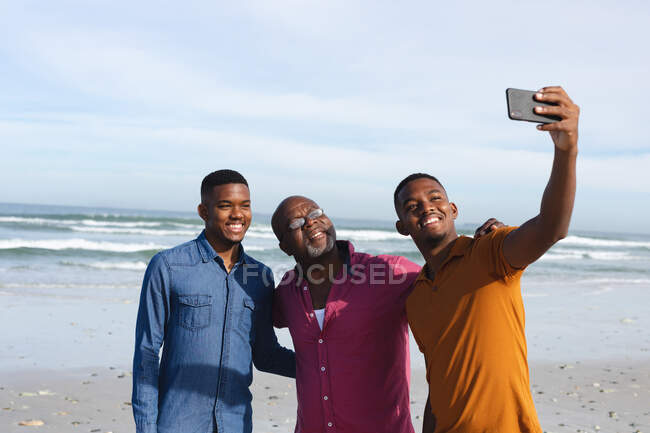 Der afroamerikanische Vater und seine beiden Söhne machen am Strand ein Selfie mit dem Smartphone. Sommer-Strandurlaub und Freizeitkonzept. — Stockfoto