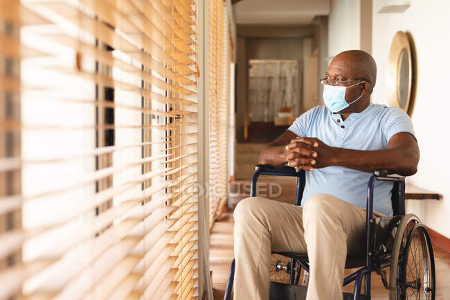 Homme âgé afro-américain handicapé portant un masque assis sur le fauteuil roulant à la maison. retraite et solitude des personnes âgées concept — Photo de stock