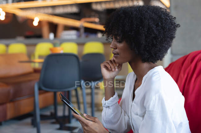 Donna afroamericana seduta sul pouf, usando tablet nel caffè. creativi digitali in movimento. — Foto stock