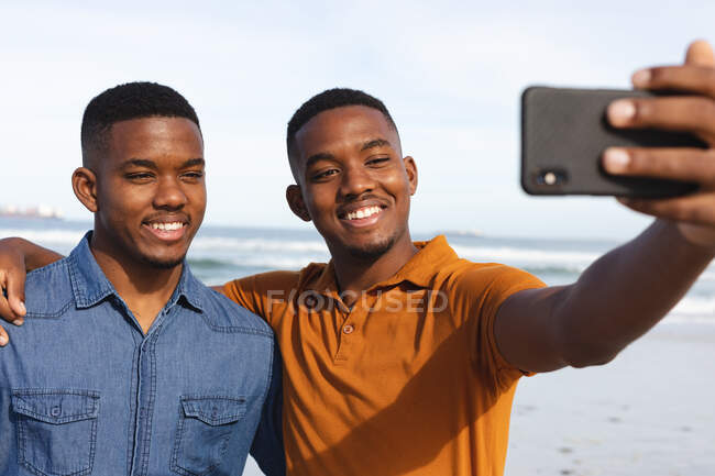 African american brothers smiling while taking a selfie from smartphone at the beach. summer beach holiday and leisure concept. — Stock Photo