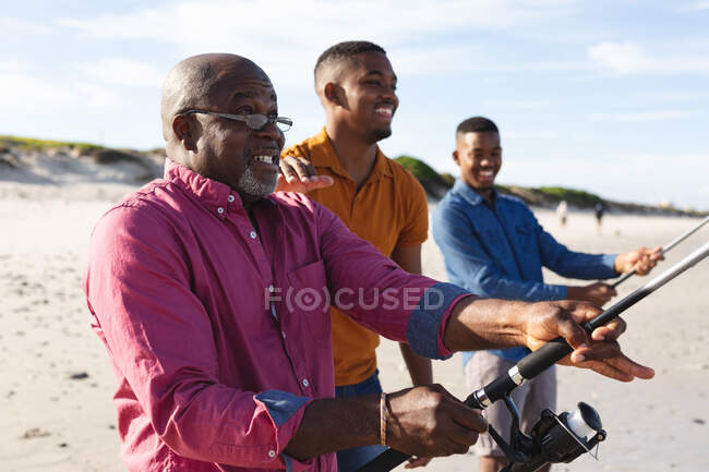 Père afro-américain et ses deux fils avec cannes à pêche pêchant ensemble à la plage. vacances à la plage d'été et concept loisirs. — Photo de stock