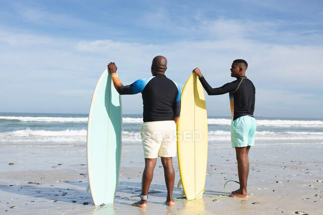 Padre e hijo afroamericanos con tablas de surf en la playa. vacaciones de playa de verano y concepto de ocio. - foto de stock