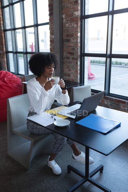 Mujer afroamericana bebiendo café, usando laptop y trabajando en la cafetería. creativos digitales sobre la marcha. - foto de stock