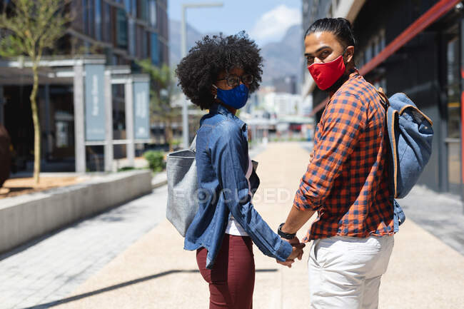 Hombre de raza mixta y mujer afroamericana usando máscaras, tomados de la mano. pasando el rato juntos durante la pandemia del coronavirus covid 19. - foto de stock