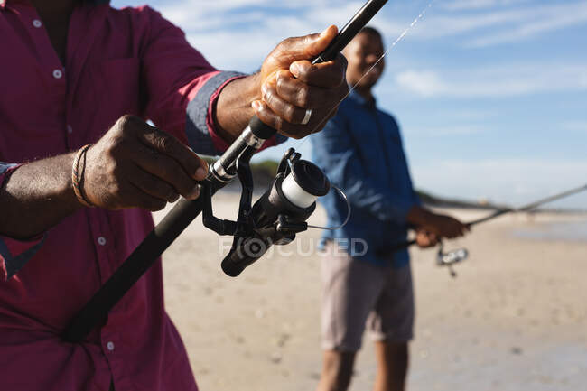 Parte média do pai afro-americano e seu filho com varas de pesca pescando juntos na praia. verão praia férias e lazer conceito. — Fotografia de Stock