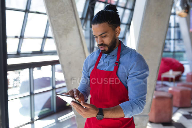 Misto raça barista masculino no avental usando tablet no café. café independente, pequeno negócio bem sucedido. — Fotografia de Stock