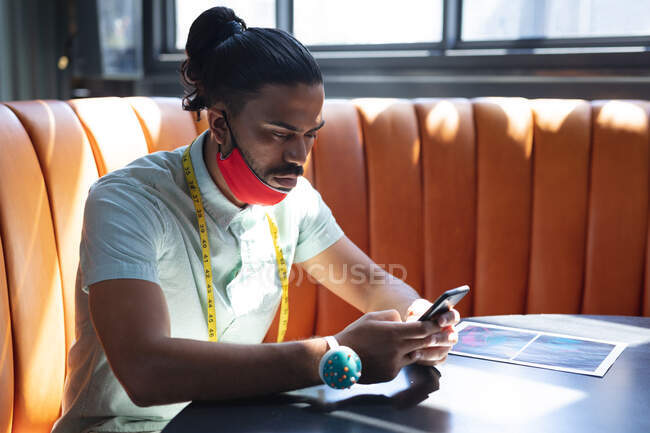 Mixed race male designer wearing face mask and using smartphone. independent creative fashion design business during coronavirus covid — Stock Photo