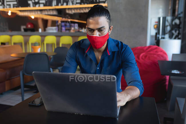 Смешанный гонщик в маске, сидит, пользуется ноутбуком в кафе. Цифровые креативы на ходу во время пандемии коронавируса ковид 19. — стоковое фото