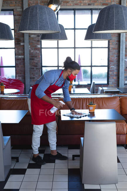 Camarero masculino de raza mixta usando mascarilla facial, desinfectando mesas en la cafetería. café independiente, negocio durante coronavirus covid 19 pandemia. - foto de stock