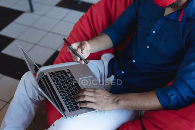 Milieu de l'homme de race mixte portant un masque facial, assis, en utilisant un smartphone et un ordinateur portable. créatifs numériques sur la route pendant coronavirus covid 19 pandémie. — Photo de stock