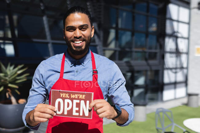 Happy mixed race male waiter holding open sign and looking at camera. independent cafe, small successful business. — Stock Photo