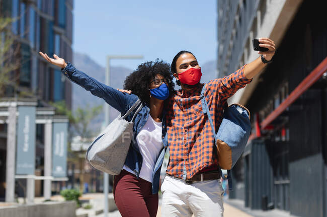 Hombre de raza mixta y mujer afroamericana usando máscaras, abrazando, tomando selfie. pasando el rato juntos durante la pandemia del coronavirus covid 19. - foto de stock