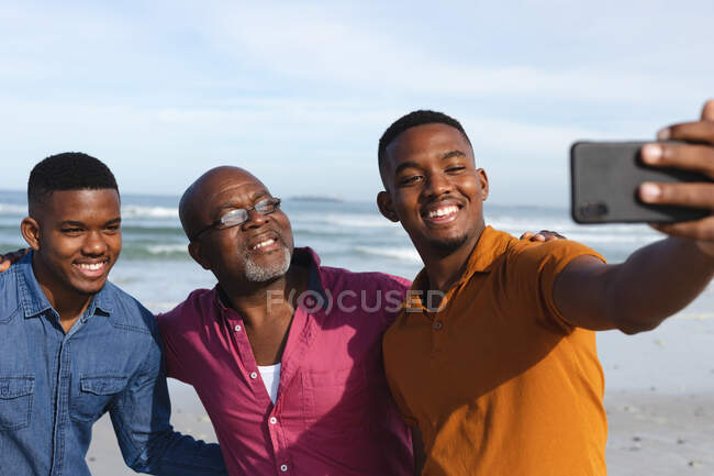 Le père afro-américain et ses deux fils prennent un selfie sur un smartphone à la plage. vacances à la plage d'été et concept loisirs. — Photo de stock