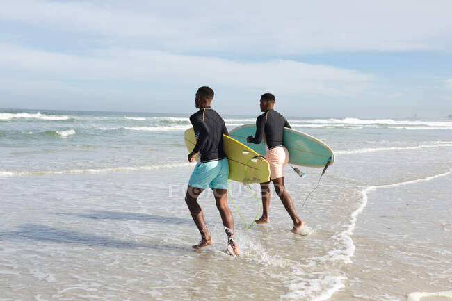Afroamerikanische Brüder mit Surfbrettern rennen am Strand auf die Wellen zu. Sommer-Strandurlaub und Freizeitkonzept. — Stockfoto