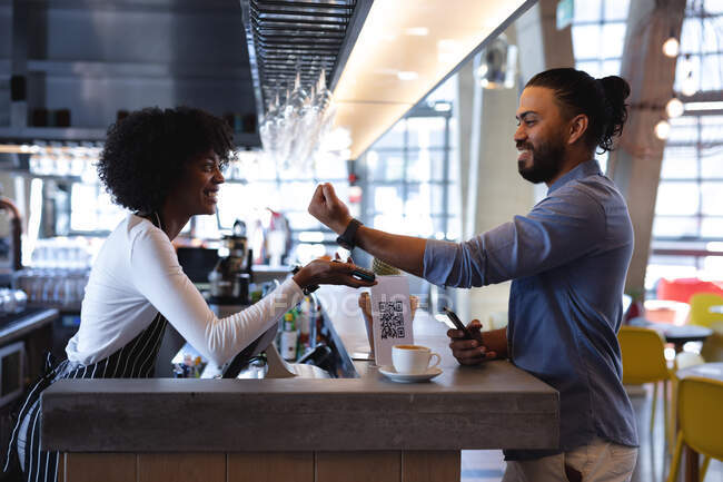 Cliente masculino de raça mista sorrindo pagando com smartwatch para barista americana africana. café independente, pequeno negócio bem sucedido. — Fotografia de Stock