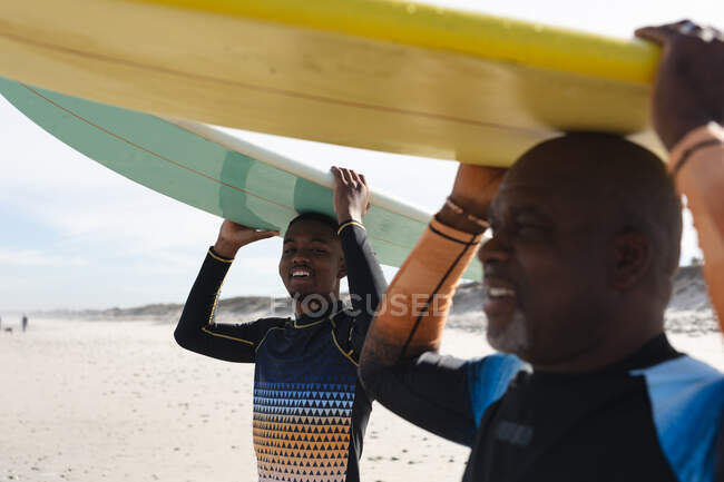 Padre e hijo afroamericanos llevando tablas de surf en sus cabezas en la playa. vacaciones de playa de verano y concepto de ocio. - foto de stock