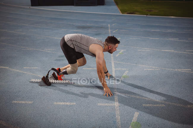 Atleta masculino caucásico con pierna protésica en posición inicial para correr en pista por la noche. concepto de deporte paralímpico - foto de stock