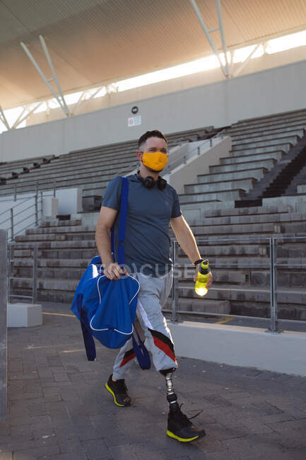 Atleta masculino caucásico con pierna protésica con máscara facial caminando en el estadio. deporte paralímpico y concepto epidémico de covid-19 - foto de stock