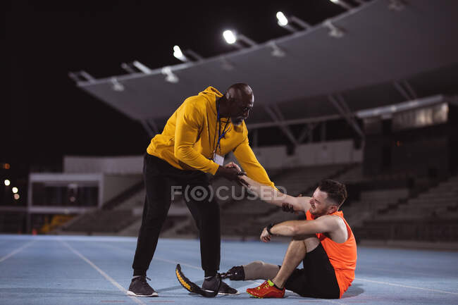 Afrikanisch-amerikanischer Männertrainer hilft kaukasischen männlichen Athleten mit Beinprothesen, auf die Beine zu kommen. Konzept des paralympischen Sports — Stockfoto