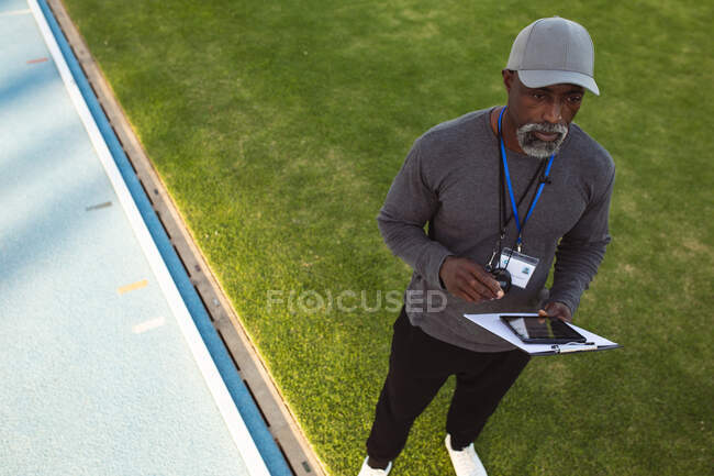 Treinador masculino afro-americano com prancheta e tablet digital em pé perto da pista de corrida. conceito de esporte paralímpico — Fotografia de Stock
