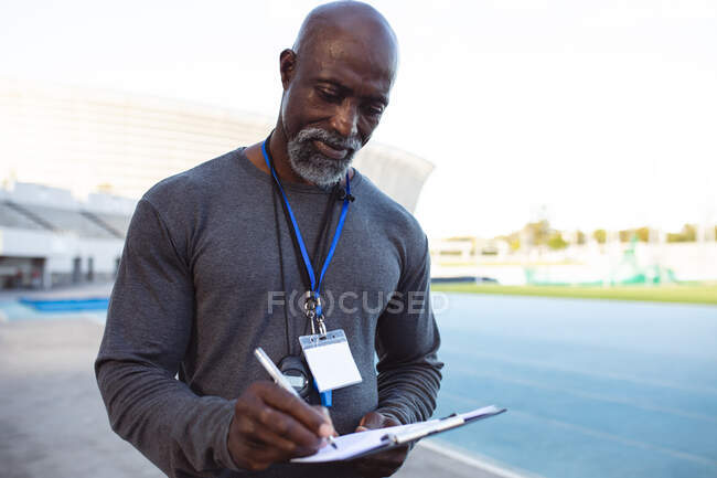 Afrikanischer Trainer mit Klemmbrett, der Notizen macht, während er im Stadion steht. Konzept des paralympischen Sports — Stockfoto