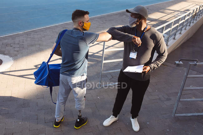 Caucasian male athlete with prosthetic leg and african american coach touching elbows in the stadium. paralympic sport and covid-19 epidemic concept — Stock Photo