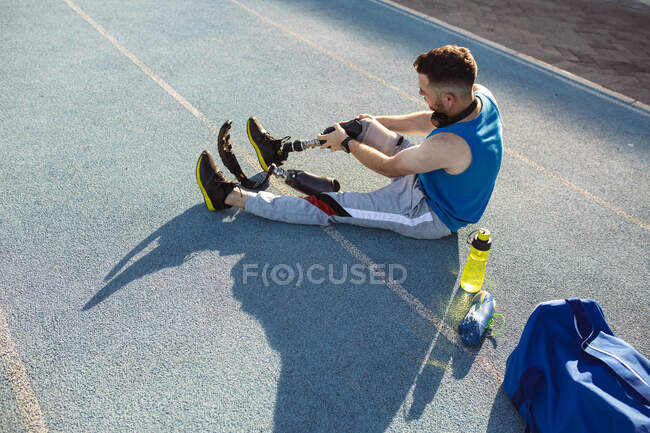 Athlète masculin caucasien fixant sa jambe prothétique tout en étant assis sur la piste de course dans le stade. concept de sport paralympique — Photo de stock