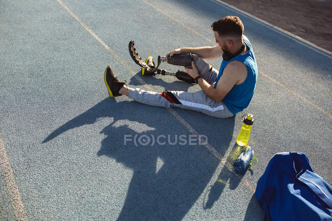 Athlète masculin caucasien fixant sa jambe prothétique tout en étant assis sur la piste de course dans le stade. concept de sport paralympique — Photo de stock