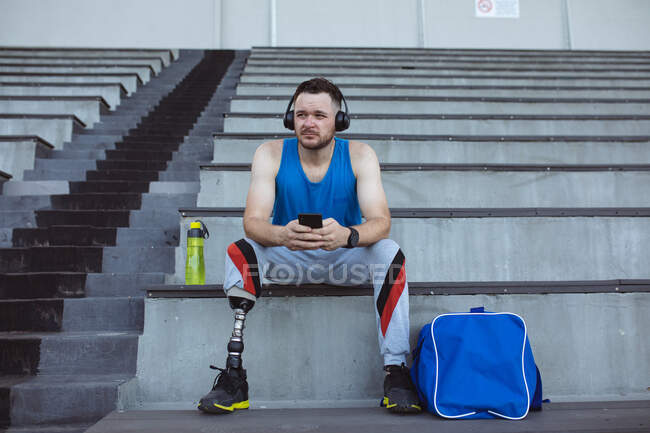 Atleta masculino caucasiano com perna protética usando smartphone sentado nos assentos no estádio. conceito de esporte paralímpico — Fotografia de Stock