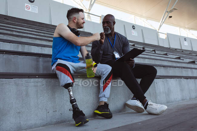 African american male coach and caucasian male athlete with prosthetic leg fist bumping in stadium. paralympic sport concept — Stock Photo