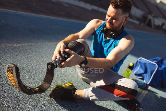 Atleta masculino caucásico arreglando su pierna protésica mientras está sentado en pista de atletismo en el estadio. concepto de deporte paralímpico - foto de stock