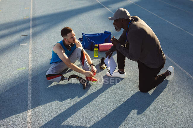 Entrenador masculino afroamericano instruyendo a atleta masculino caucásico con pierna protésica en el estadio. concepto de deporte paralímpico - foto de stock