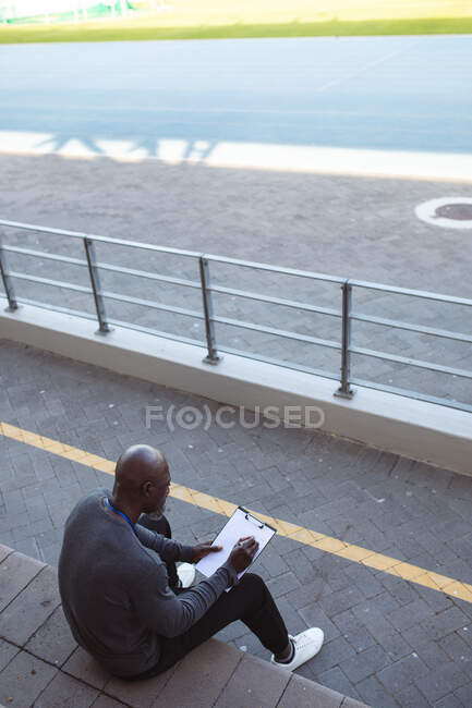 Entrenador masculino afroamericano con portapapeles tomando notas mientras está sentado en los asientos del estadio. concepto de deporte paralímpico - foto de stock