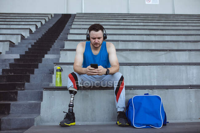 Atleta masculino caucasiano com perna protética usando smartphone sentado nos assentos no estádio. conceito de esporte paralímpico — Fotografia de Stock
