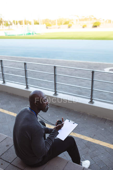 Treinador masculino afro-americano com cronômetro medindo tempo enquanto sentado nos assentos no estádio. conceito de esporte paralímpico — Fotografia de Stock