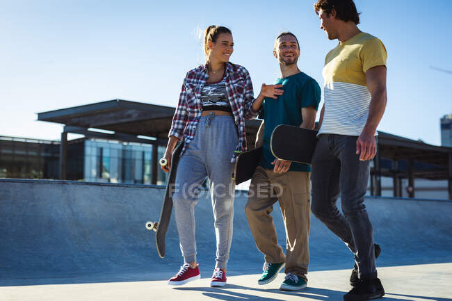 Трое счастливых кавказских женщин и мужчин ходят со скейтбордами и разговаривают на солнце. тусоваться в городском скейтпарке летом. — стоковое фото