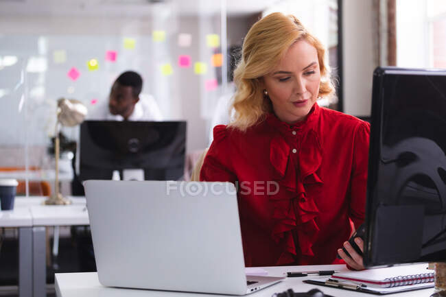 Kaukasische Frau mit Smartphone, während sie auf ihrem Schreibtisch im modernen Büro sitzt. Business, Professionalität und Bürokonzept — Stockfoto